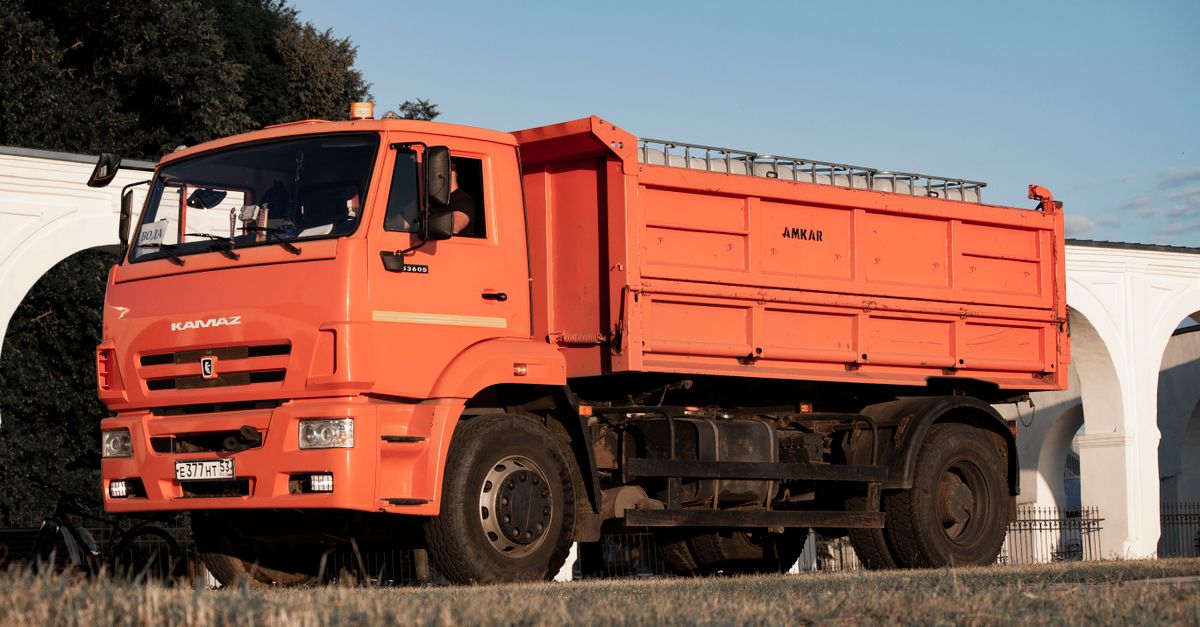 An orange KAMAZ truck positioned outdoors against a backdrop of arches and greenery.